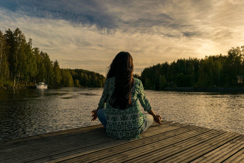 Eine Frau im Meditations-Sitz auf einem Holzsteg mit Blick auf einen See.