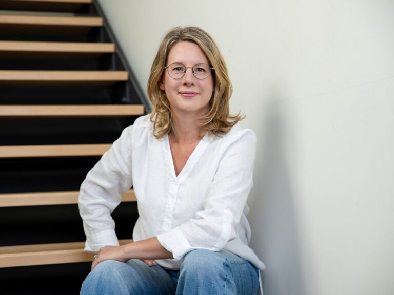 Stefanie Kappus sitzt auf einer Holztreppe vor einer weißen Wand, den linken Ellbogen auf ihr Knie gestützt, mit offenem und freundlichem Blick in die Kamera.