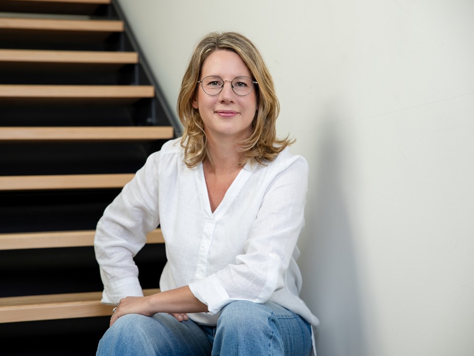 Stefanie Kappus sitzt auf einer Holztreppe vor einer weißen Wand, den linken Ellbogen auf ihr Knie gestützt, mit offenem und freundlichem Blick in die Kamera.